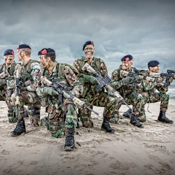 Sex soldater i kamouflage och baskrar, som håller gevär, knäböjer på en sandstrand med havet i bakgrunden under en molnig himmel.