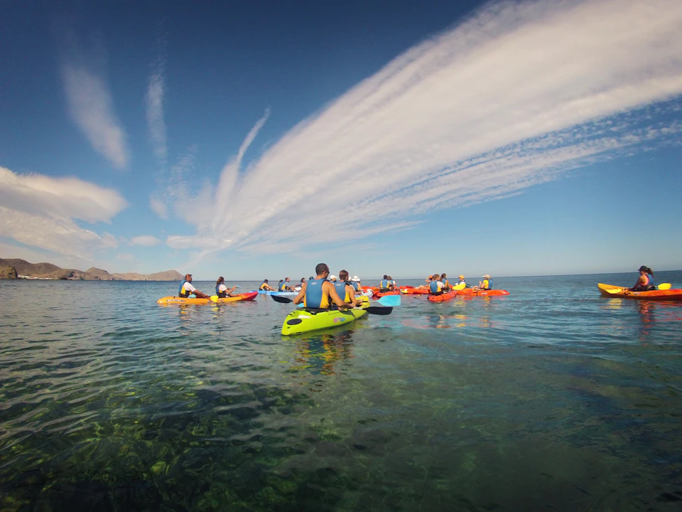 People kayaking on clear, calm waters beneath a blue sky streaked with clouds, with distant mountains visible in the background.