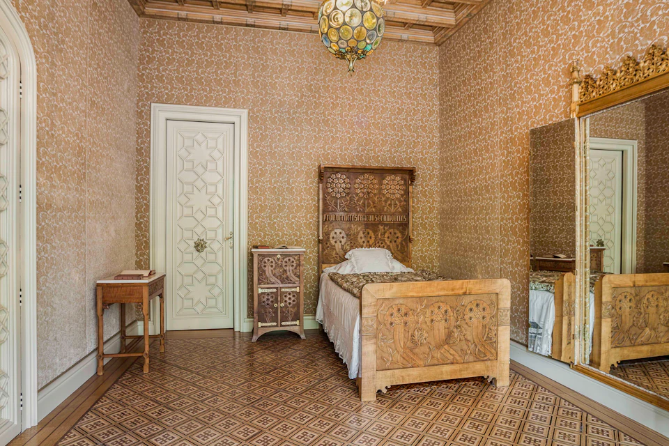 Ornate bedroom with patterned wallpaper, a wooden bed, nightstand, small table, mirrored wardrobe, and tiled floor.