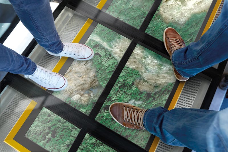 Due paia di piedi in scarpe da ginnastica poggiano su un pavimento di vetro che rivela una vista su un paesaggio boscoso sottostante.