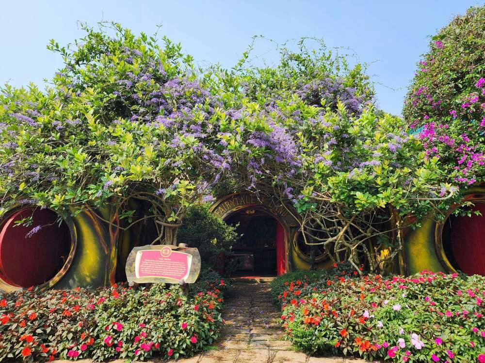 A whimsical garden entrance covered in purple flowers and greenery, with colorful flowers and a decorative sign in front.