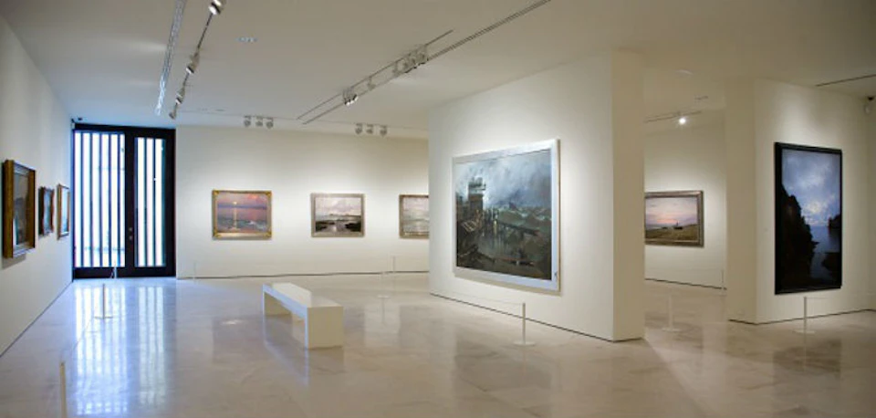 Bright art gallery with landscape paintings on white walls, a central bench, and track lighting overhead.