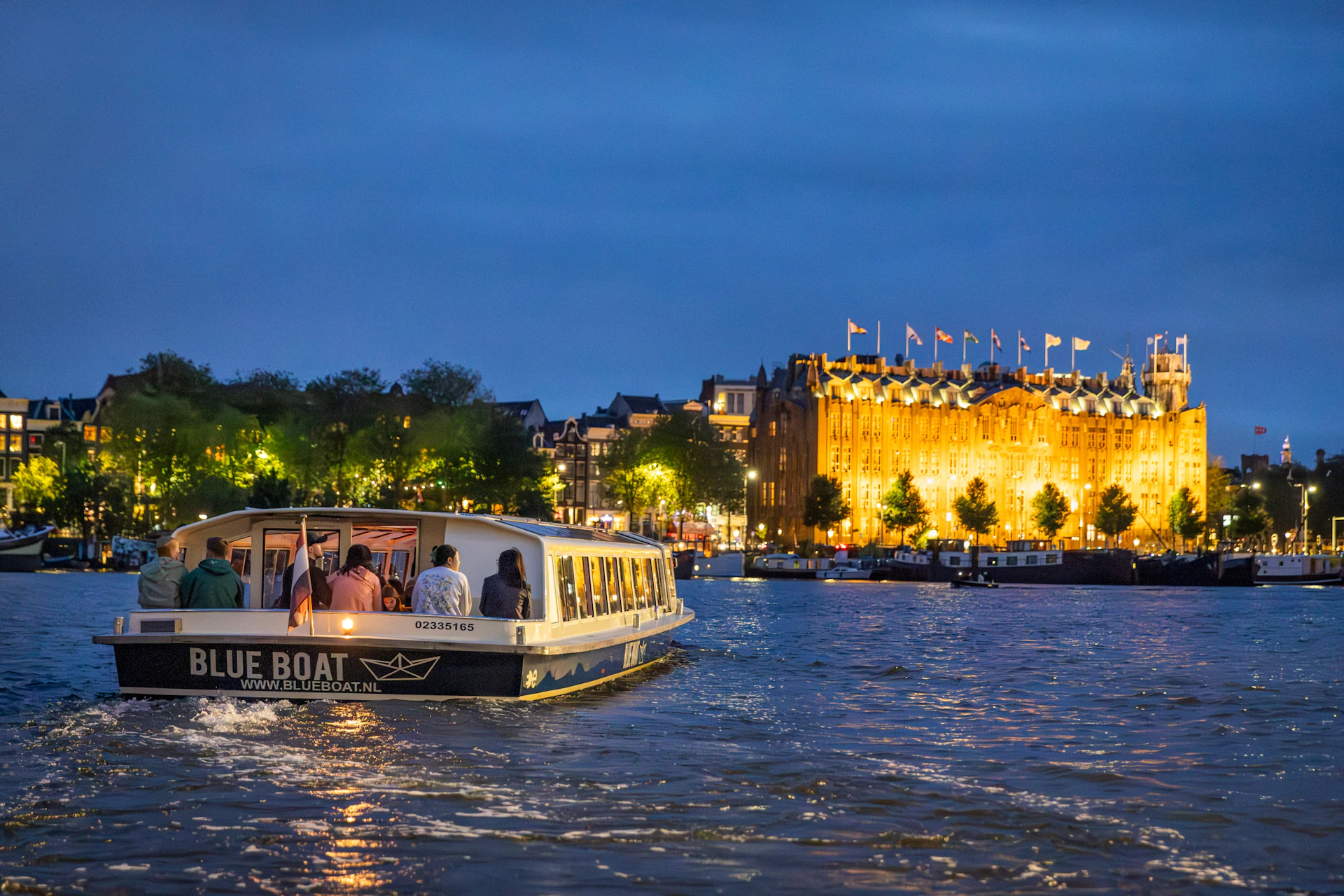 A boat with passengers glides on a river at night, passing a brightly lit building with flags on its roof.