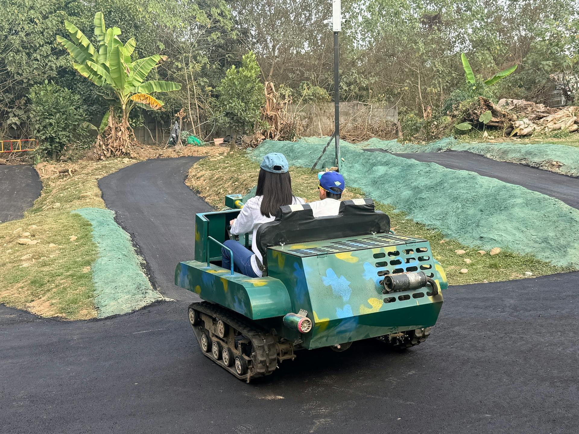 Two people ride a small camouflaged tank-like vehicle on a paved path surrounded by greenery.