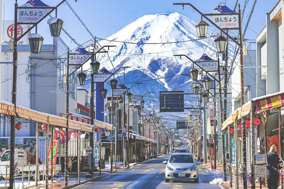 A street lined with shops and streetlights, leading to a view of Mount Fuji under a clear blue sky.