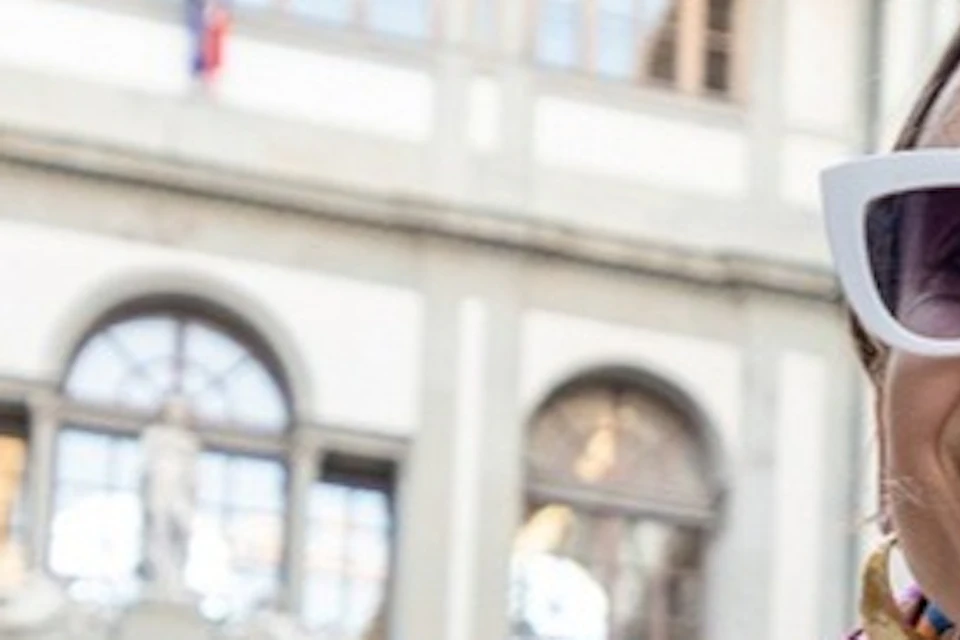 A woman wearing sunglasses, a polka-dot dress, and a scarf smiles in an open courtyard surrounded by classical architecture.