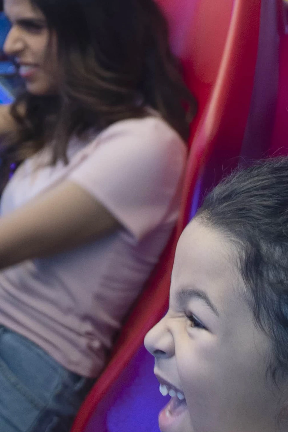 Two people excitedly play racing games on arcade driving simulators, surrounded by colorful lights and game machines.