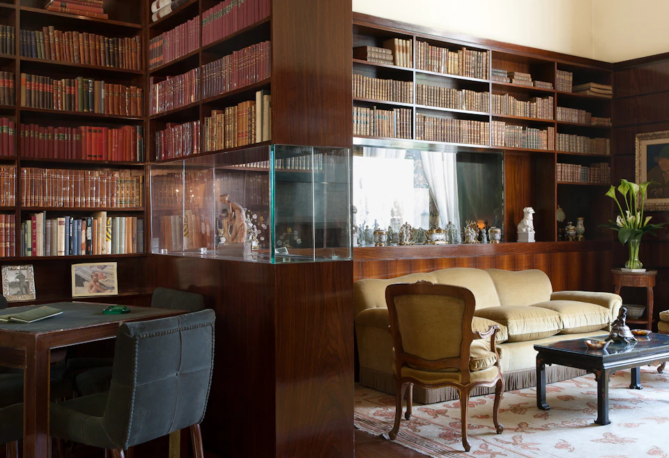 A cozy study with wooden bookshelves, antique books, a glass display case, a sofa, an armchair, and a desk with a chair.