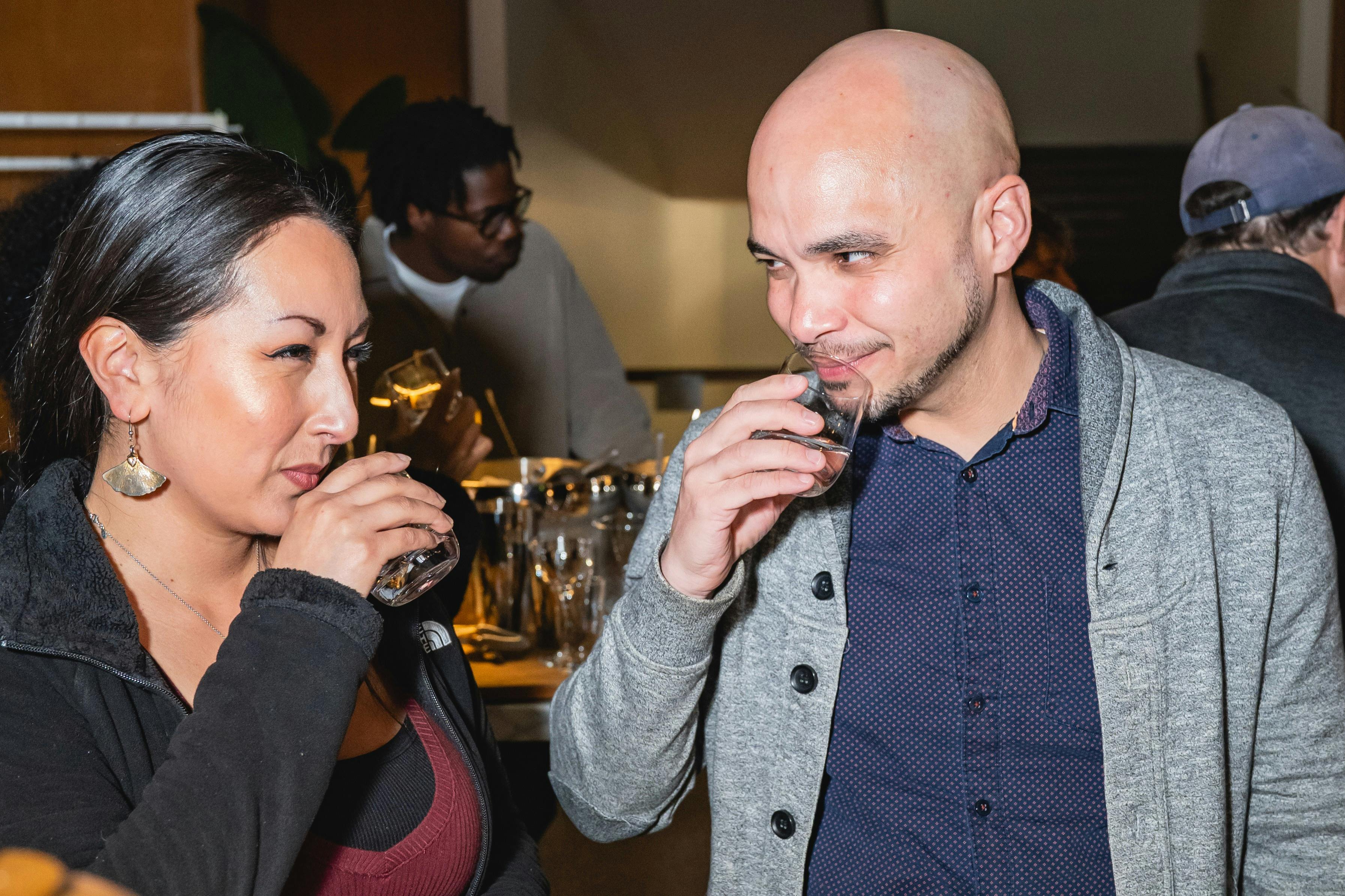 Two people in conversation holding glasses, with another person in the background in a social setting.