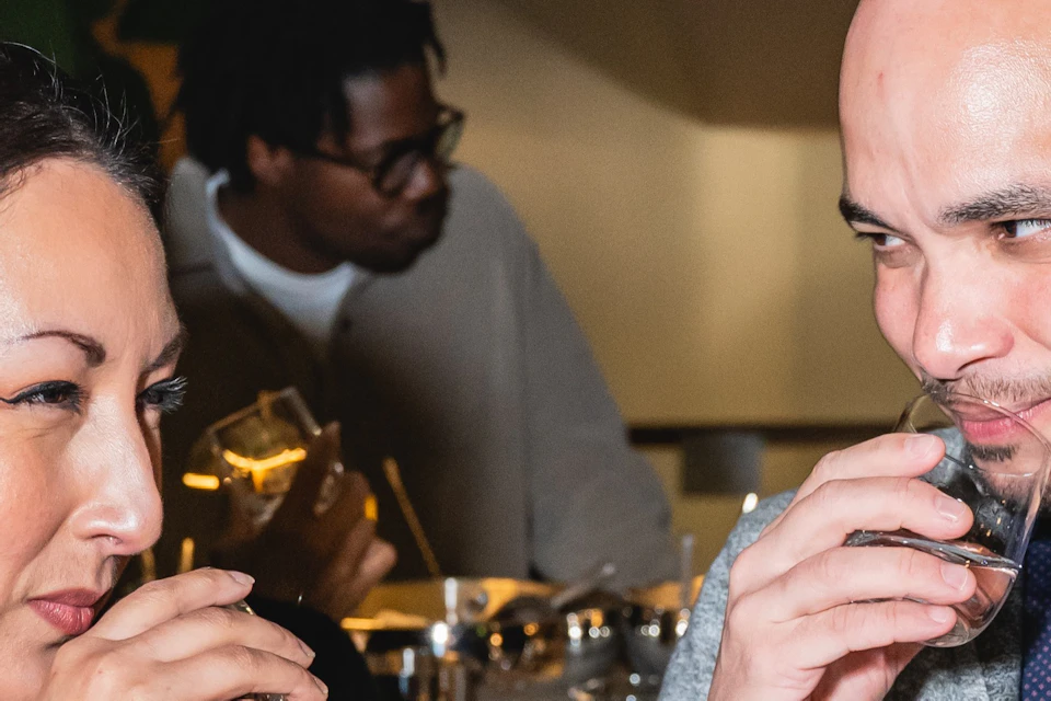 Two people in conversation holding glasses, with another person in the background in a social setting.