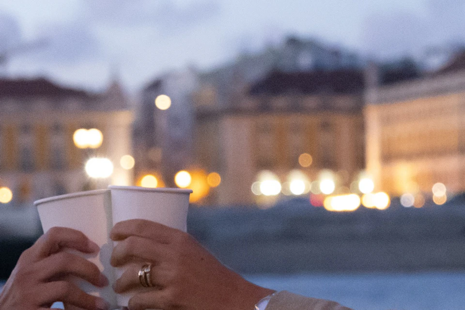 Two people clink cups on a boat in front of a lit-up cityscape at dusk. One person smiles as they toast.