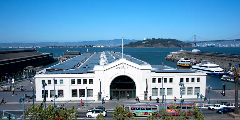 The image shows Pier 15 in San Francisco, featuring a large white building, a bay, boats, people walking, and the Bay Bridge in the background.