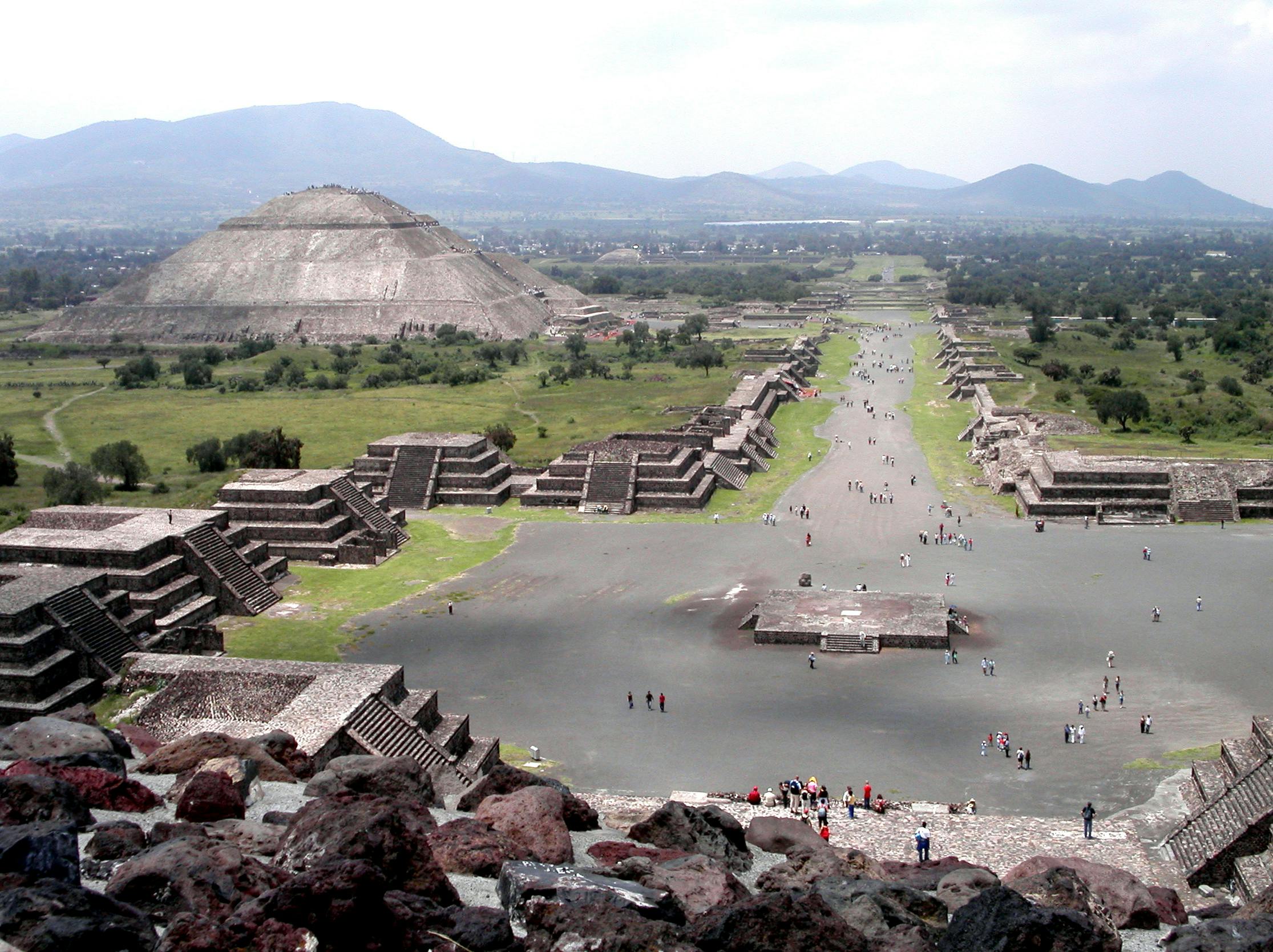 Pohled na Teotihuacán s Avenue of the Dead, pyramidami a procházejícími se lidmi. Hory jsou viditelné v pozadí.