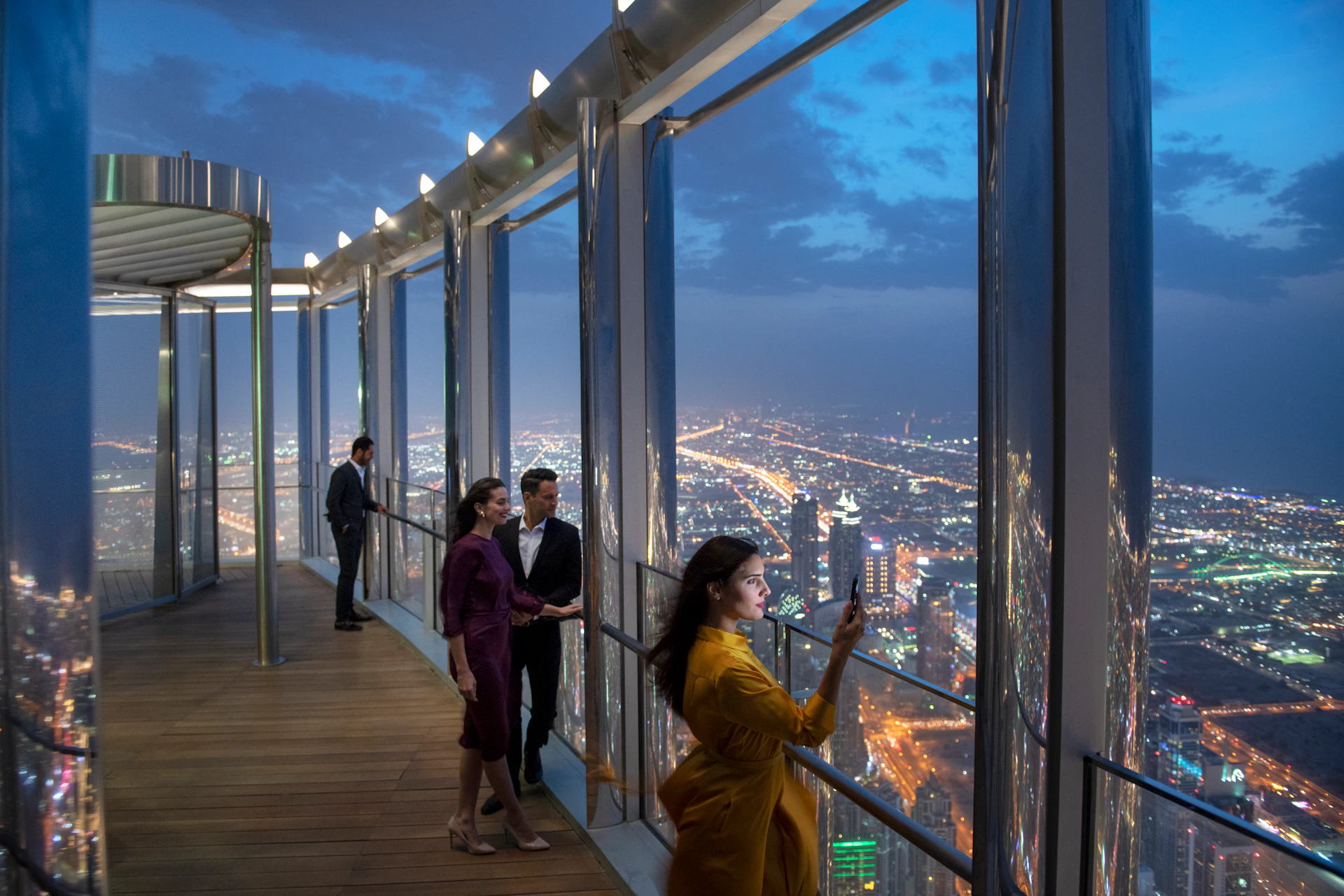 People standing on an observation deck at night, overlooking a brightly lit cityscape through large glass windows.