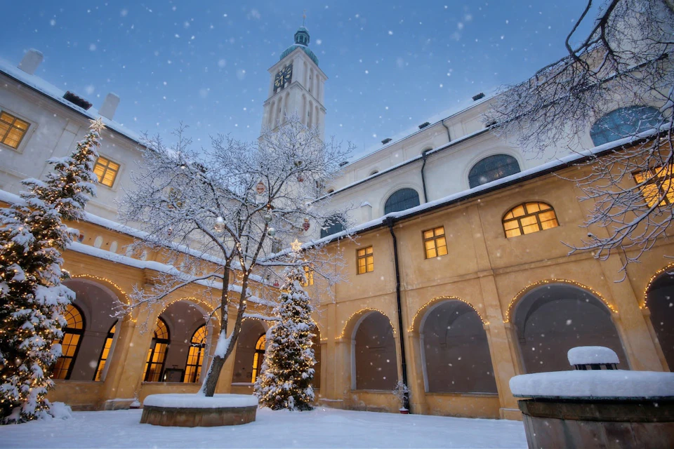 A snowy courtyard with a tree decorated for Christmas, surrounded by arched buildings and a clock tower in the background.