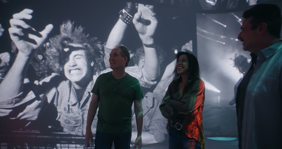 Een man in een groen shirt staat voor een grote vintage foto van enthousiaste concertbezoekers met opgeheven handen.