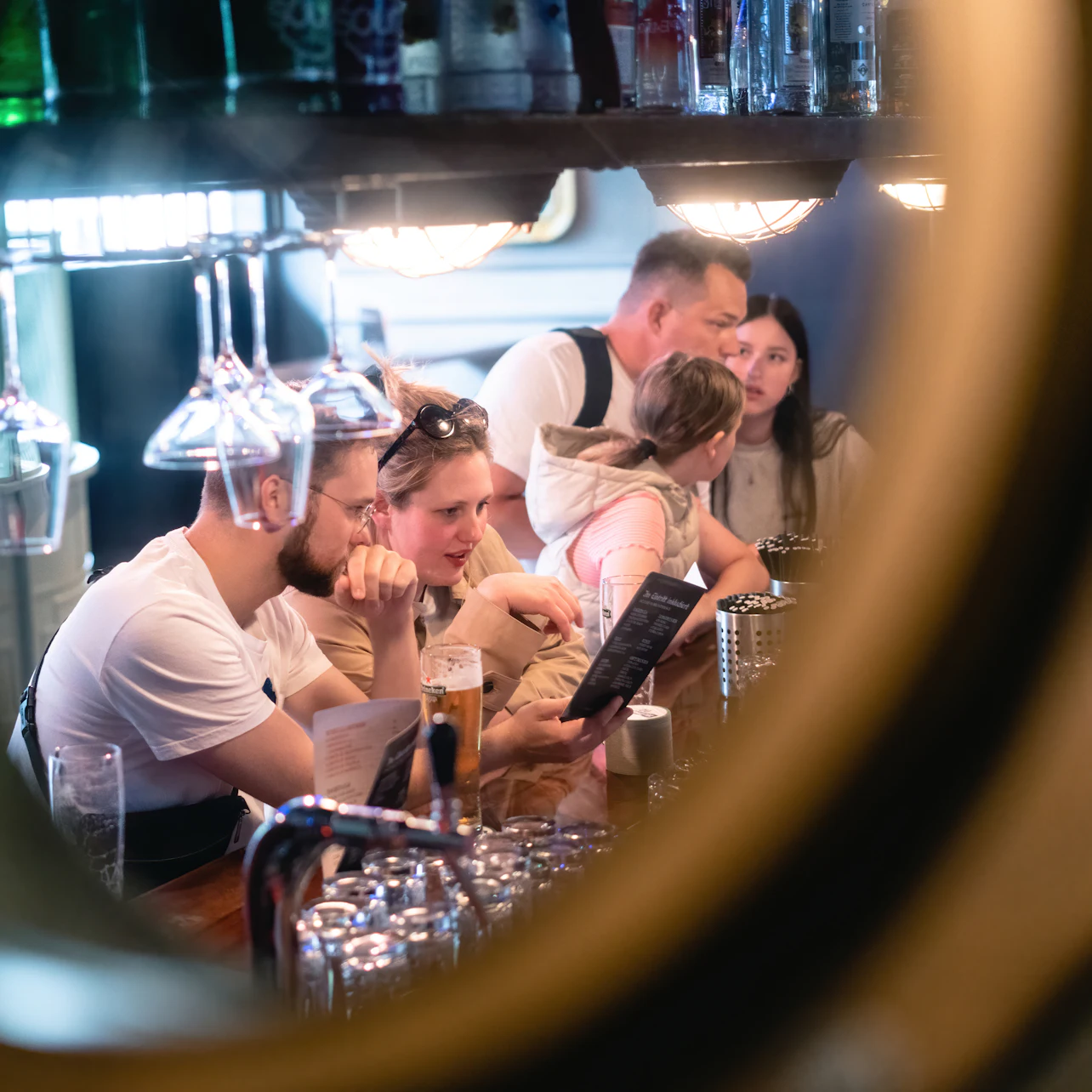 People sitting at a bar, some reading menus, with hanging glassware and shelves visible through a circular frame.