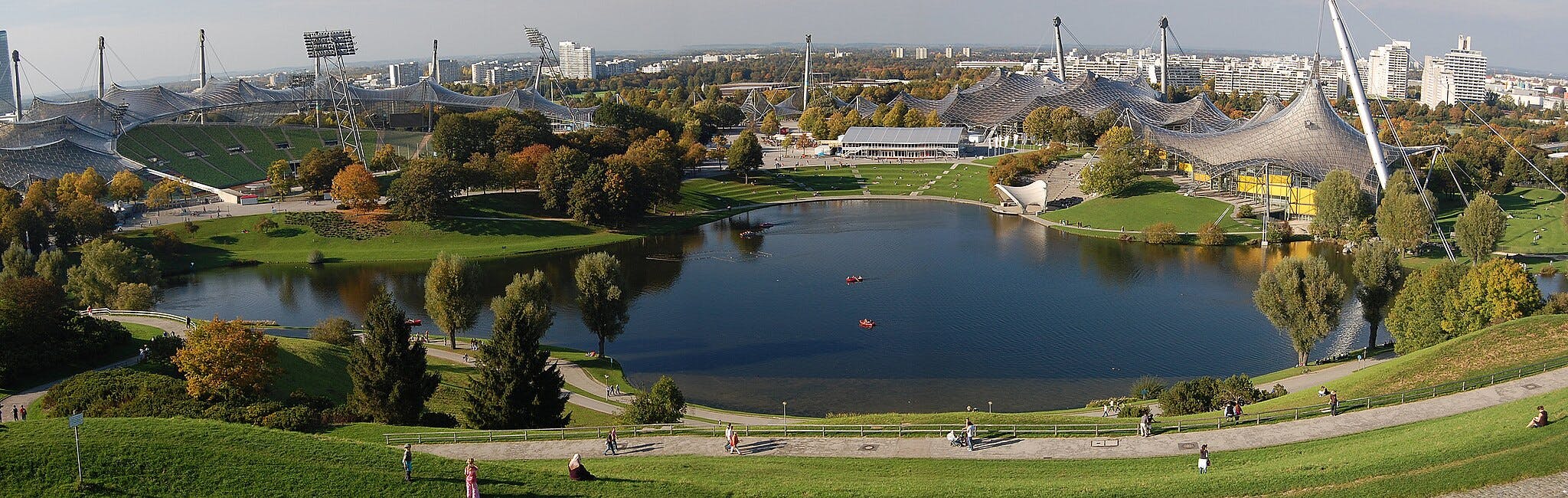 Vista dell'Olympiapark con un grande stadio, aree verdi lussureggianti, un lago con kayak ed edifici urbani sullo sfondo.