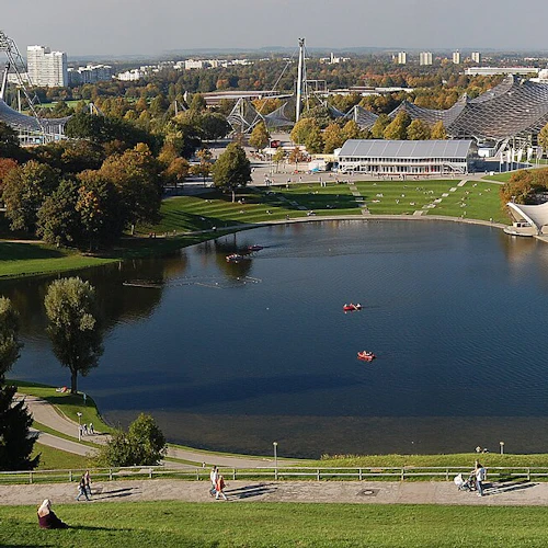 Blick auf den Olympiapark mit einem großen Stadion, üppigen Grünflächen, einem See mit Kajakfahrern und städtischen Gebäuden im Hintergrund.