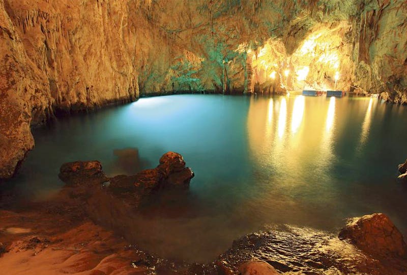 Υπόγεια πισίνα σπηλαίων με ανακλαστικό γαλάζιο νερό, που φωτίζεται από ζεστά φώτα, προβάλλοντας τραχιά τοιχώματα βράχων.
