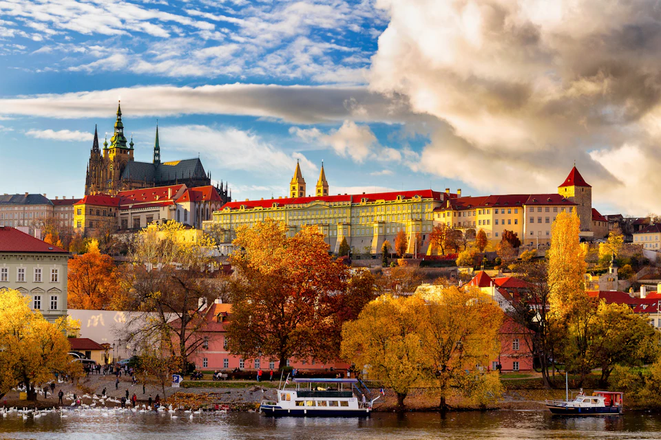Scenic view of a river with trees in autumn colors, a boat docked, historic buildings, and a cathedral under a partly cloudy sky.