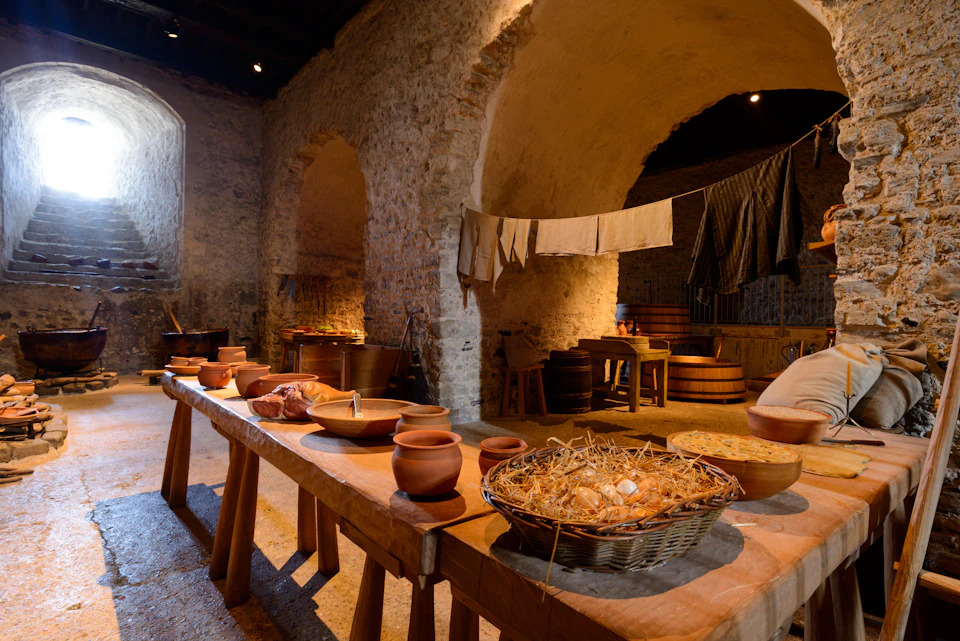 A rustic kitchen with stone walls, wooden tables, pottery, baskets of food, and cloths hanging to dry.