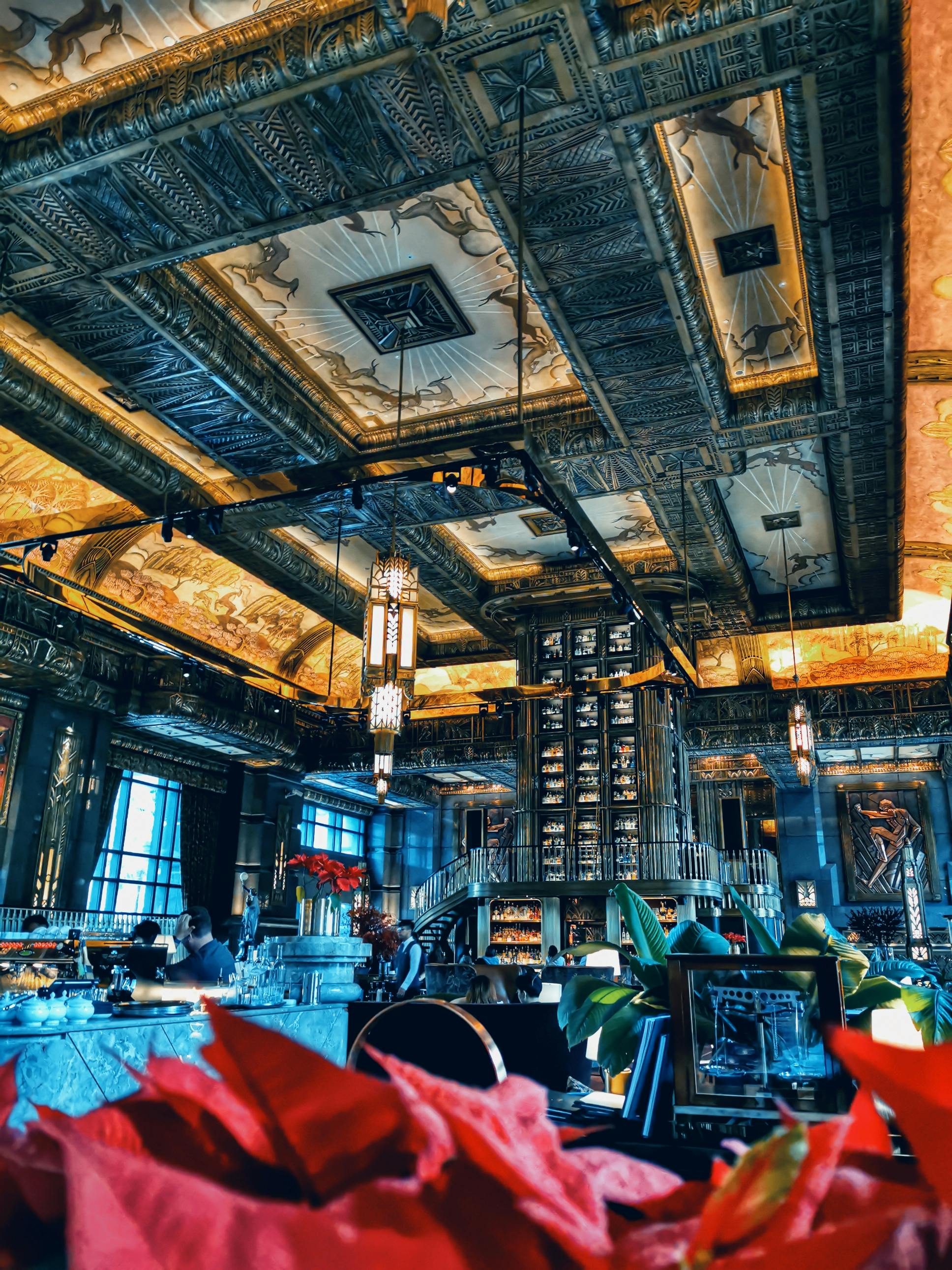 A restaurant with an ornate, golden ceiling, chandeliers, and a central bar. People sit at tables, with plants in the foreground.