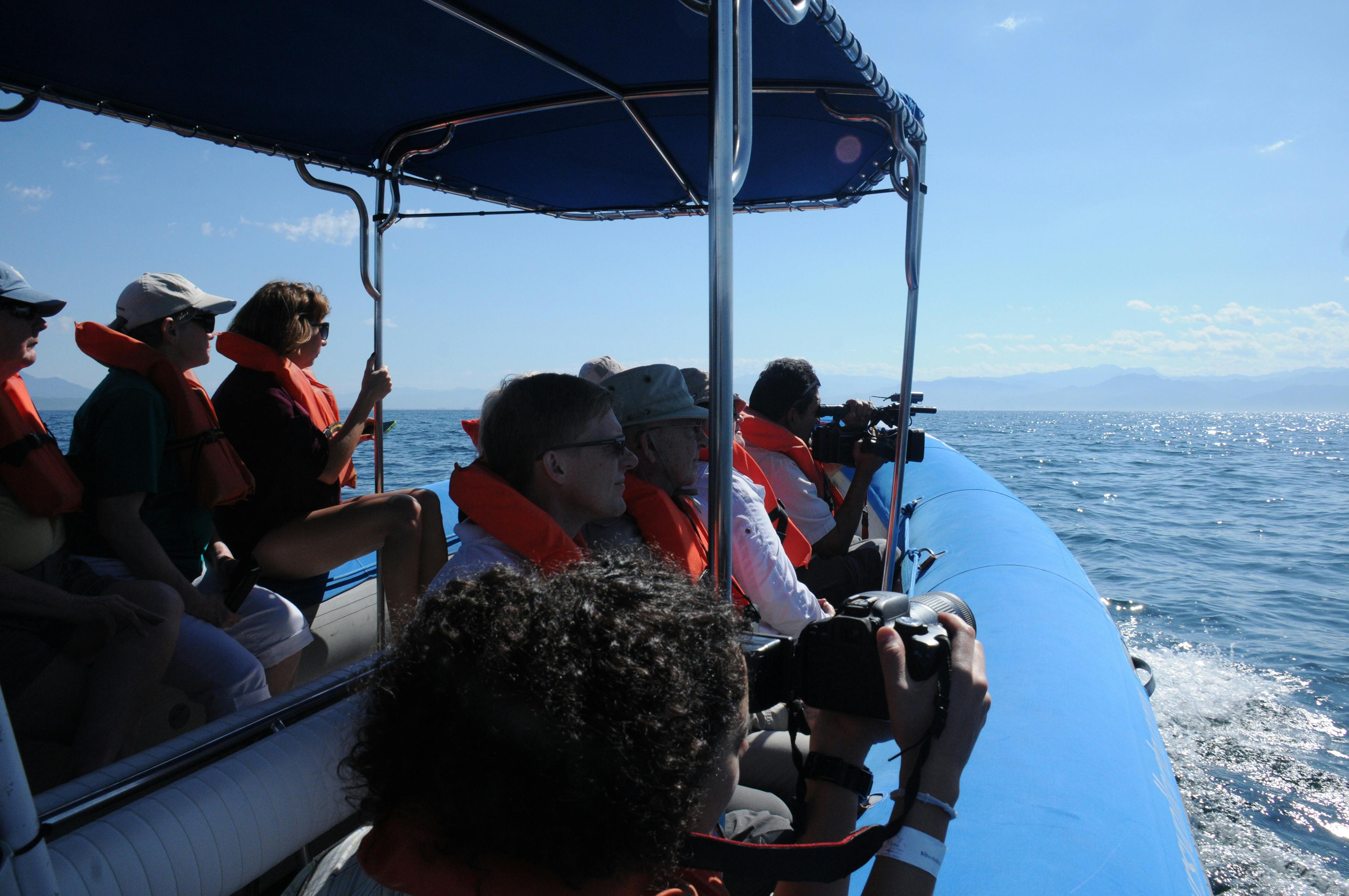 En grupp människor som bär flytvästar är på en blå kapellbåt, några tar bilder, med ett lugnt hav och avlägsna berg i sikte.