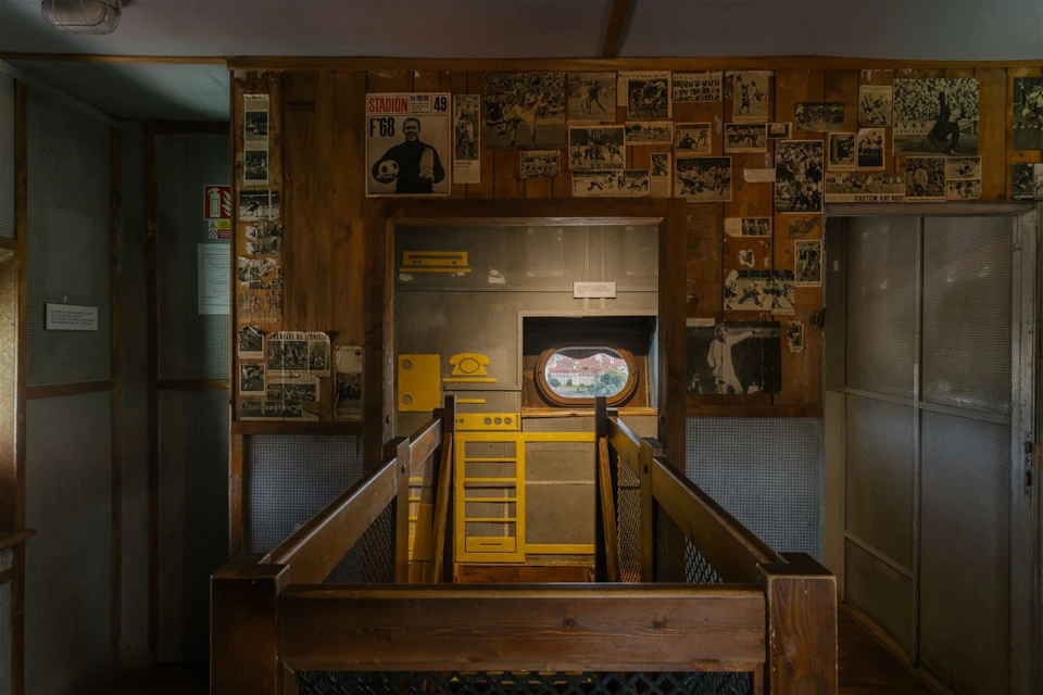 A narrow hallway with wooden railings leads to a wall decorated with vintage photos and posters. A small round window offers an outdoor view.