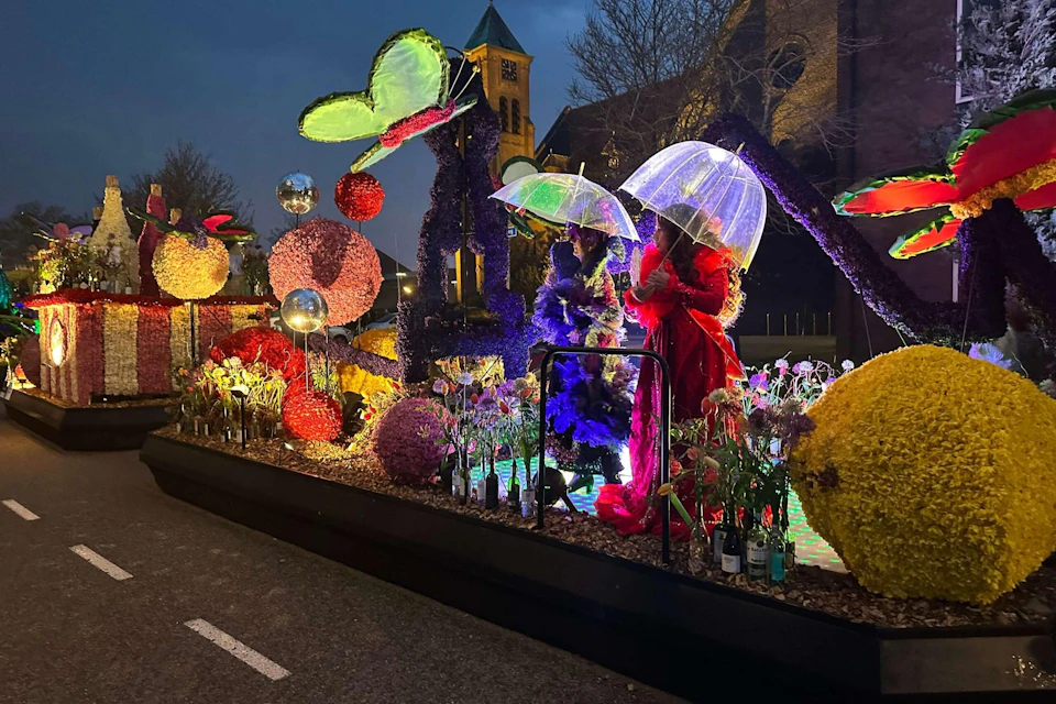 A colorful float with illuminated decorations and two people holding transparent umbrellas during a nighttime event.