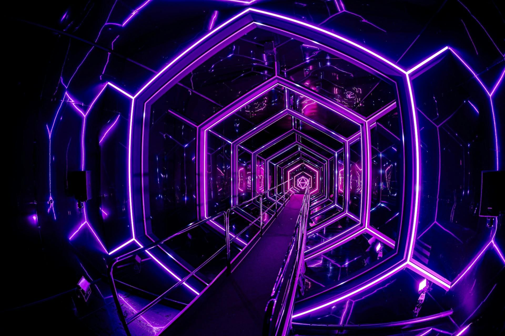 A futuristic hexagonal tunnel lit with neon purple lights, featuring a central walkway and reflective surfaces on all sides.