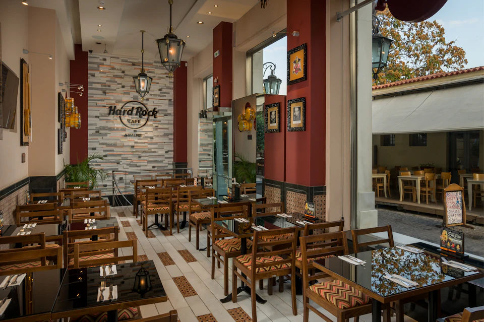 Modern restaurant interior with wooden tables and chairs, red accent walls, large windows, and a sign reading “Hard Rock Café.”