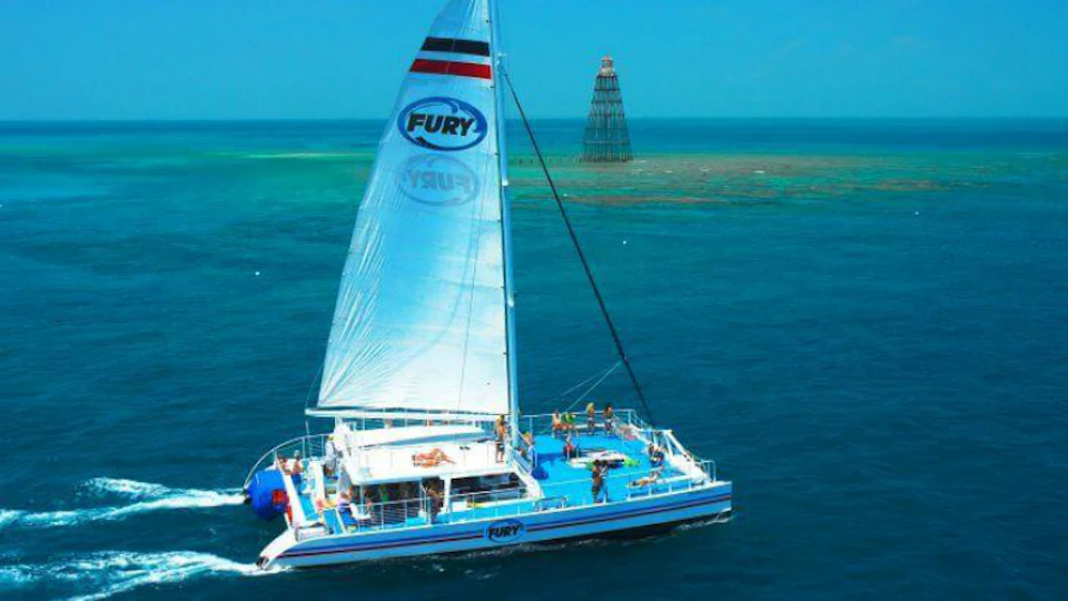A sailboat with "Fury" on the sail cruises through clear blue water, with a navigational marker structure in the distance.