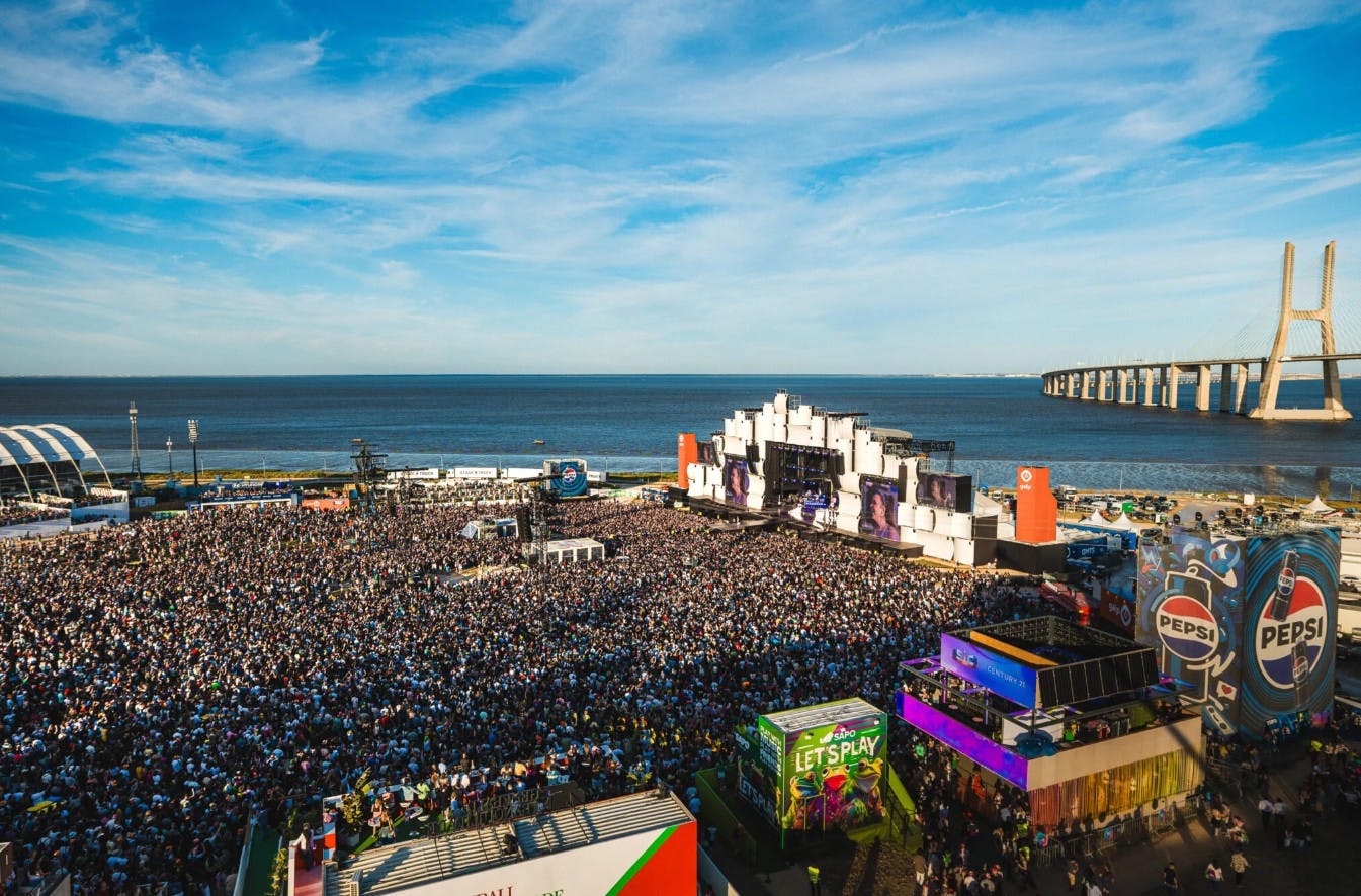 Una gran multitud de festivals de música a l'aire lliure davant d'un escenari al costat del mar sota un cel assolellat, amb estructures de colors al voltant.