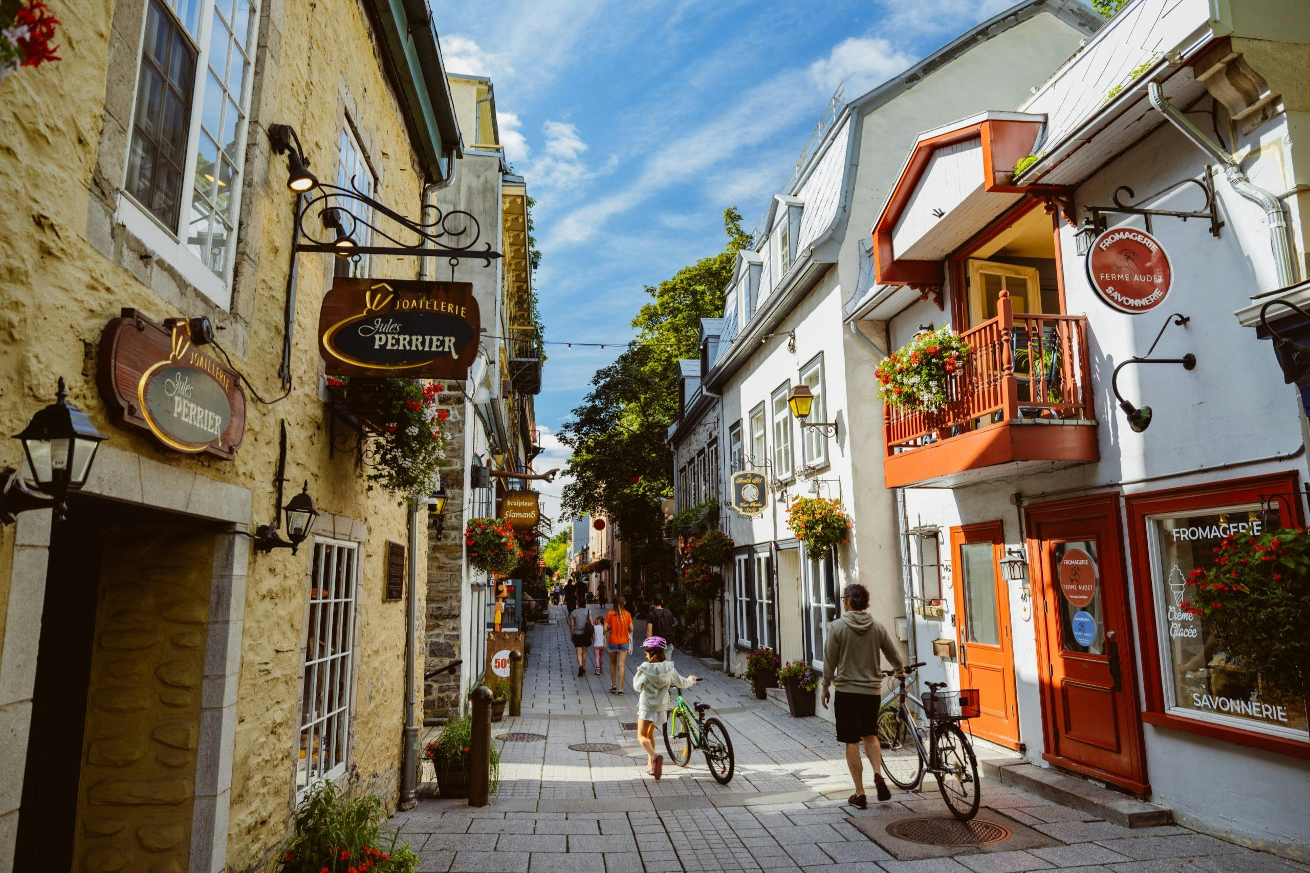 A cobblestone pedestrian street lined with shops and flowers, with people walking and biking under a bright sky.