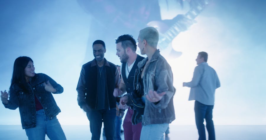 Čtyři lidé, ležérně oblečení, stojí spolu, smějí se a komunikují v dobře osvětleném, vnitřním prostředí s modře zbarveným pozadím.