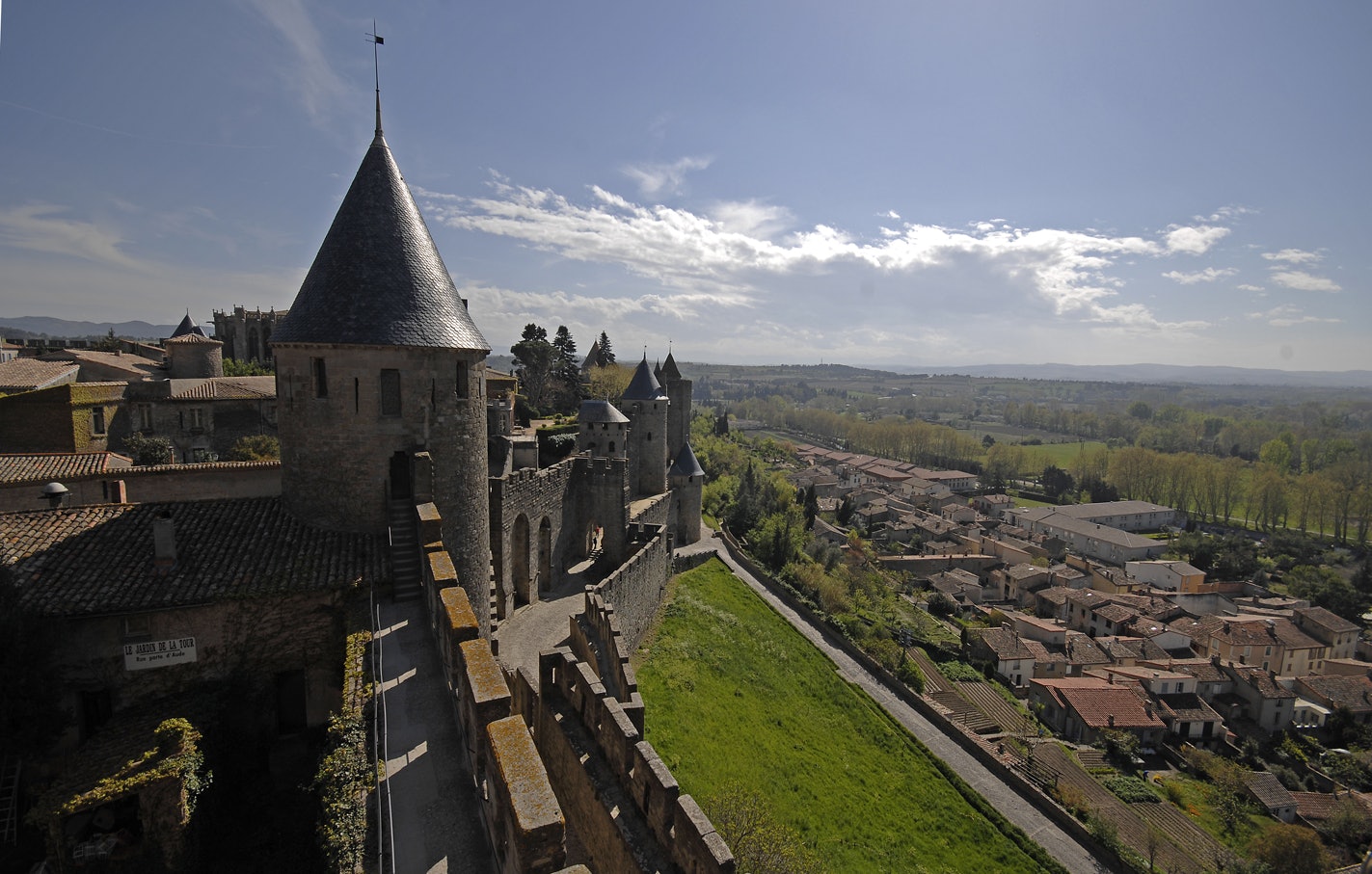 Un château historique en pierre avec des tours coniques surplombe une ville aux toits de tuiles rouges et un paysage verdoyant au loin.