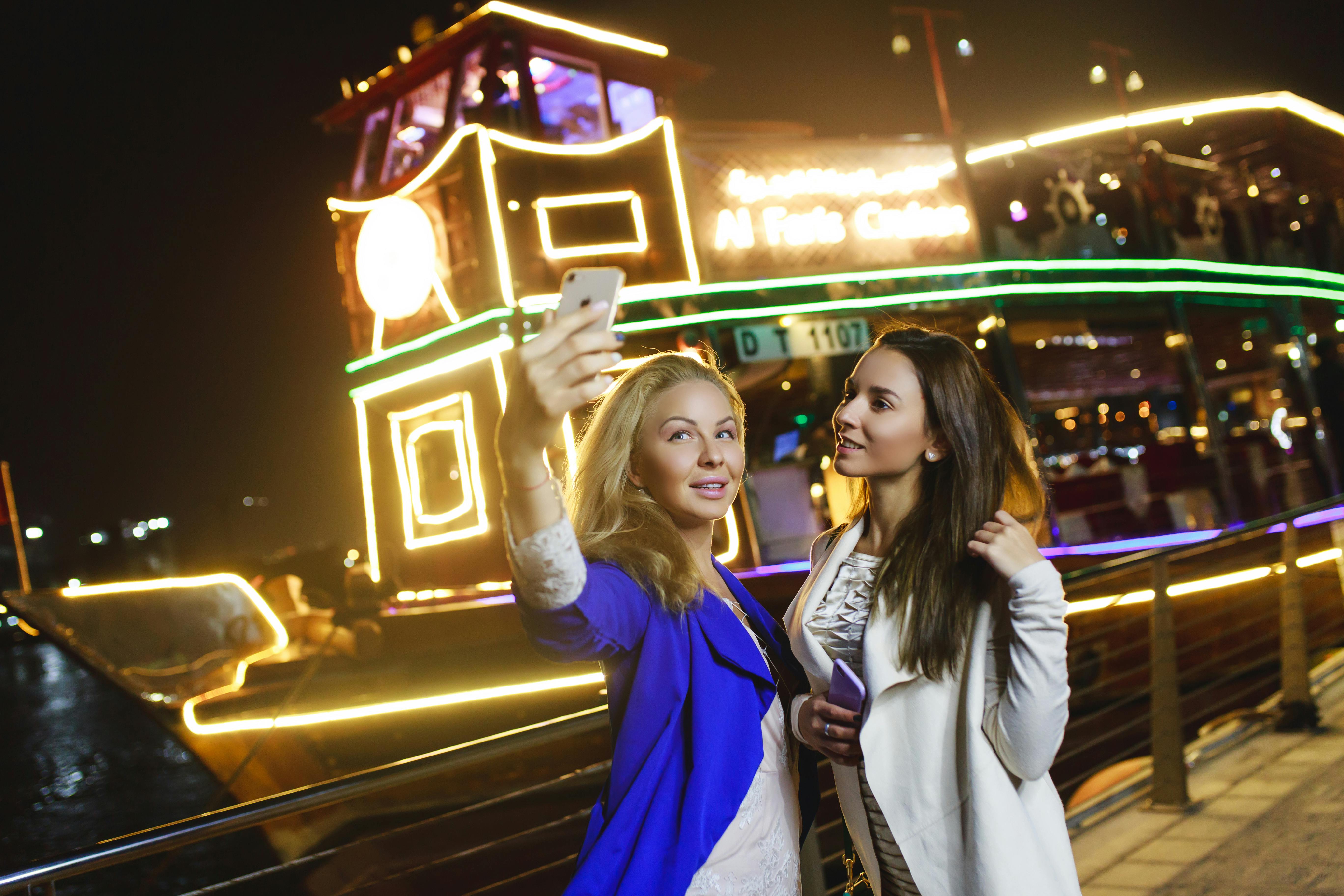 Two women taking a selfie in front of a brightly lit ship at night, with colorful lights and a festive atmosphere.