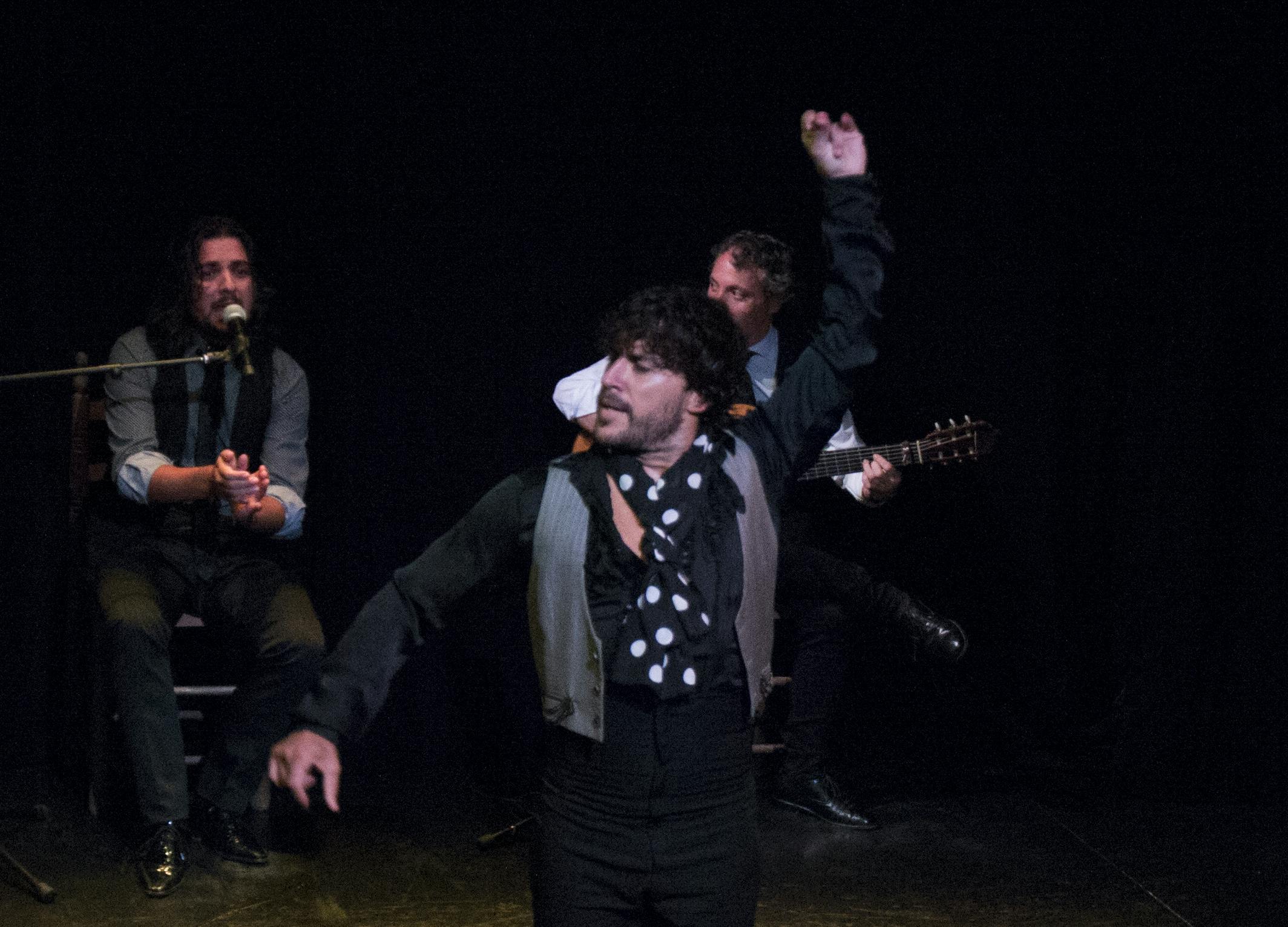 Una ballarina actua amb els braços aixecats mentre dos músics asseguts darrere d'ell canten i toquen la guitarra en un escenari poc il·luminat.