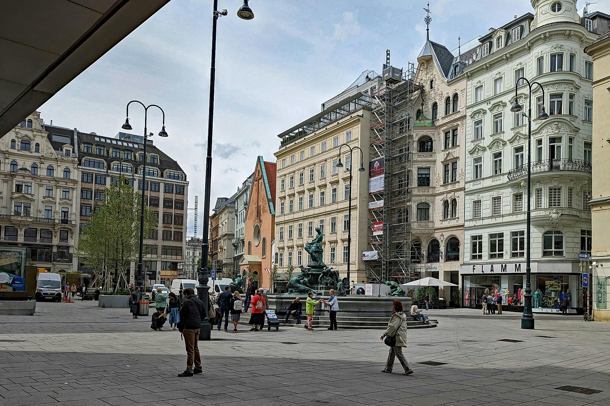 Persone che camminano e si siedono intorno a una fontana in una piazza molto frequentata, circondata da edifici storici e da una chiesa in ristrutturazione.