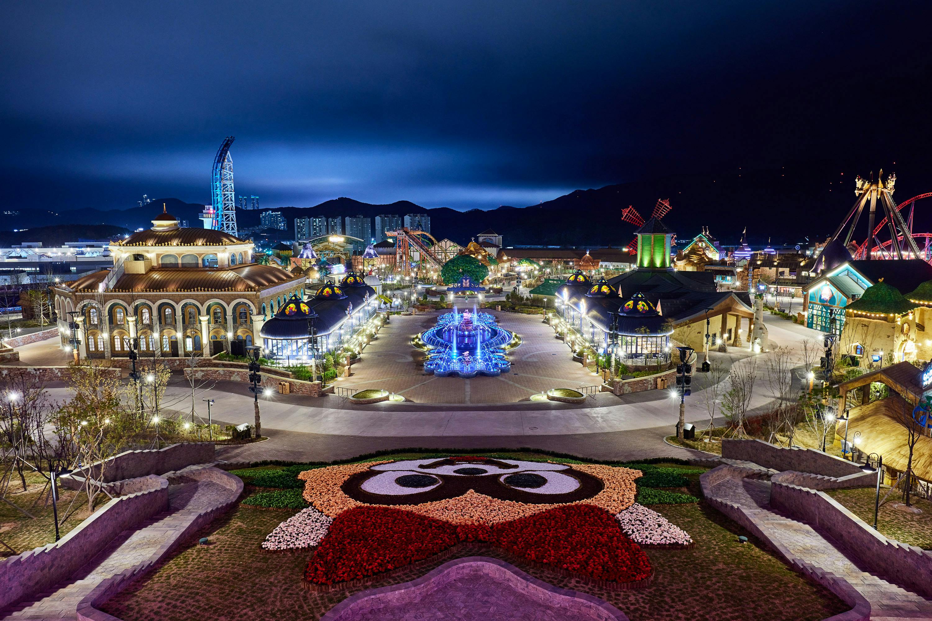 A brightly lit amusement park at night with a flower bed resembling a face in the foreground and rides in the background.