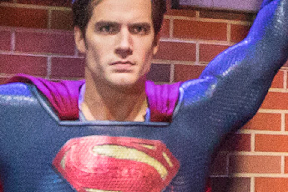 People in a room pose with a Superman statue holding up a red helicopter. The background has cityscape graphics and blue lighting.