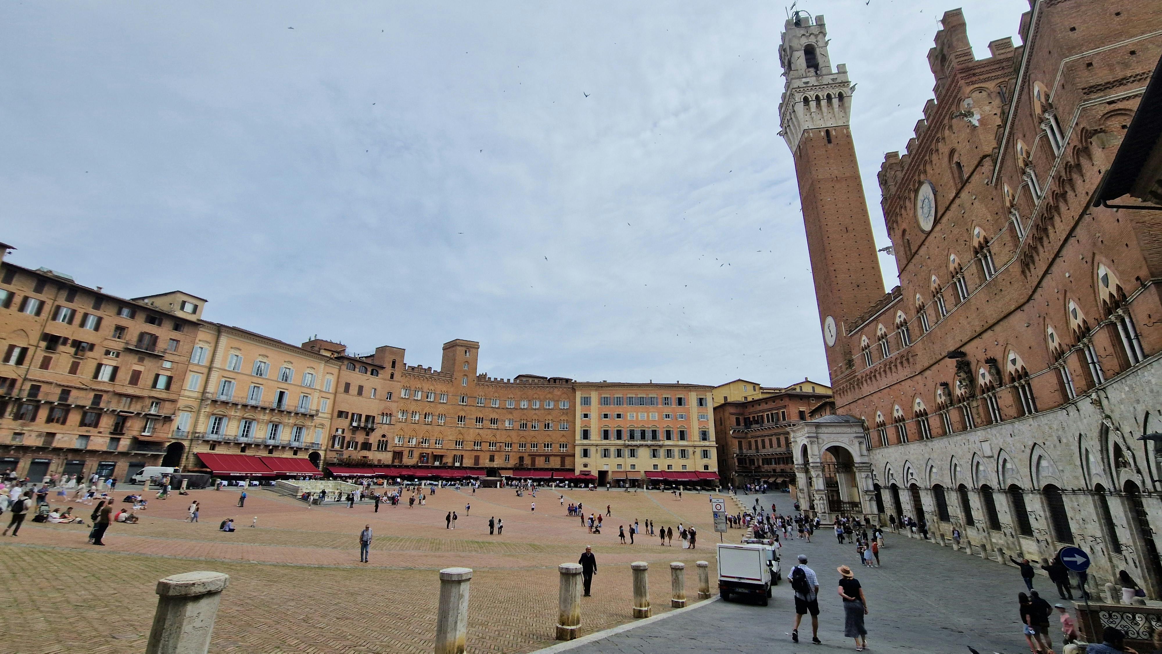 People walking in a large, open square in front of tall, historic buildings with a high tower under a partly cloudy sky.
