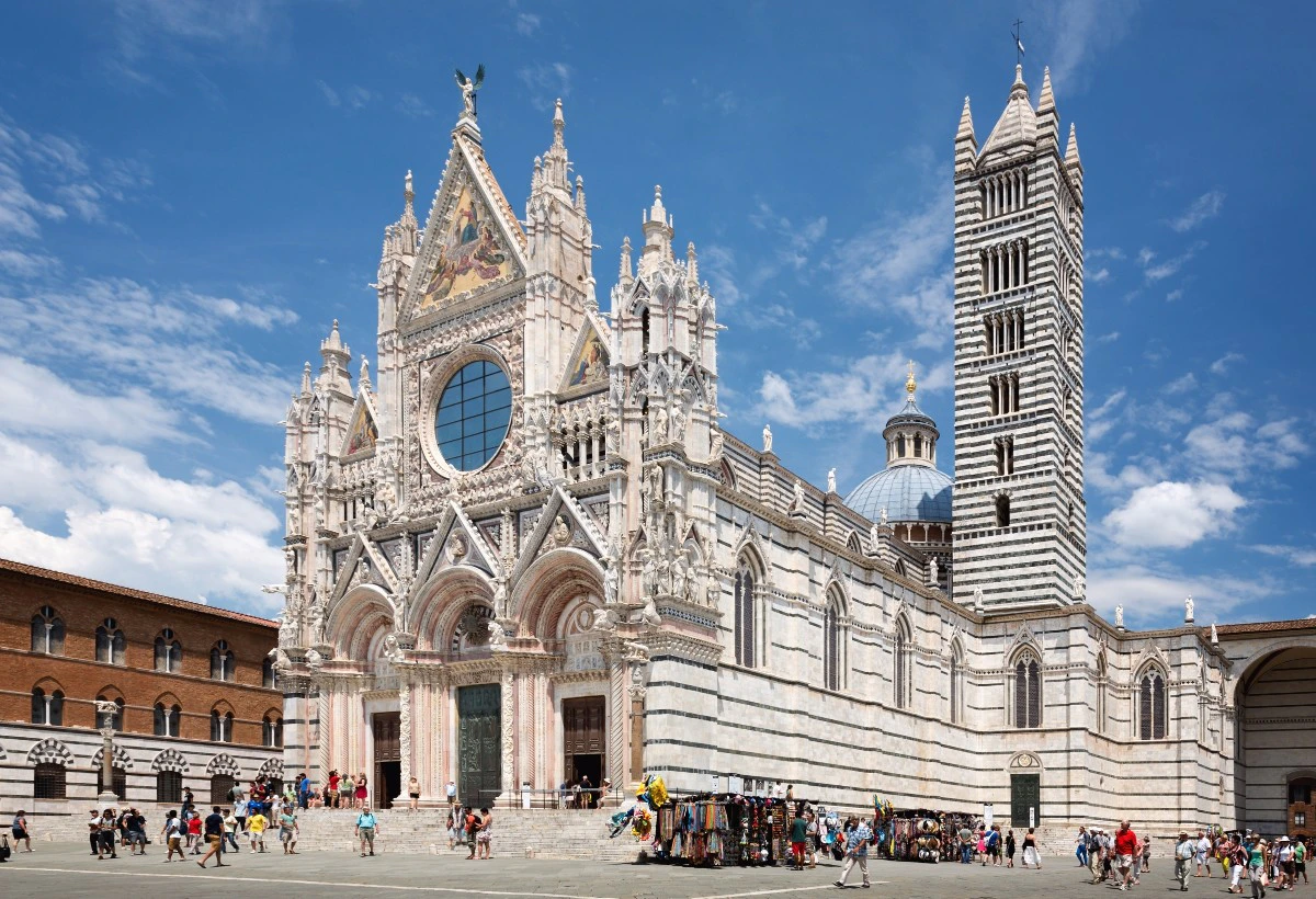 A large, ornate Gothic cathedral with a striped bell tower under a blue sky. Tourists and vendors are visible in front.