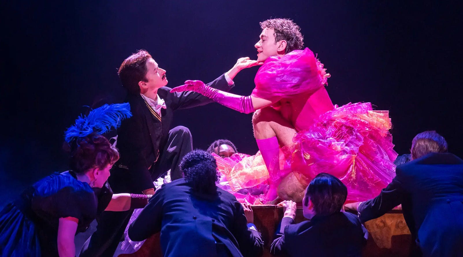 Ένα άτομο που φοράει μια φωτεινή ροζ ζωηρή φορεσιά κρατά το πηγούνι ενός άλλου ατόμου. Το φόντο απεικονίζει άλλους ντυμένους με σκούρα ρούχα.