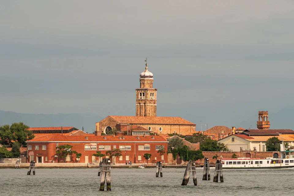 A coastal scene with a red-bricked building topped by a tall tower with a domed roof, set against a cloudy sky and calm water.
