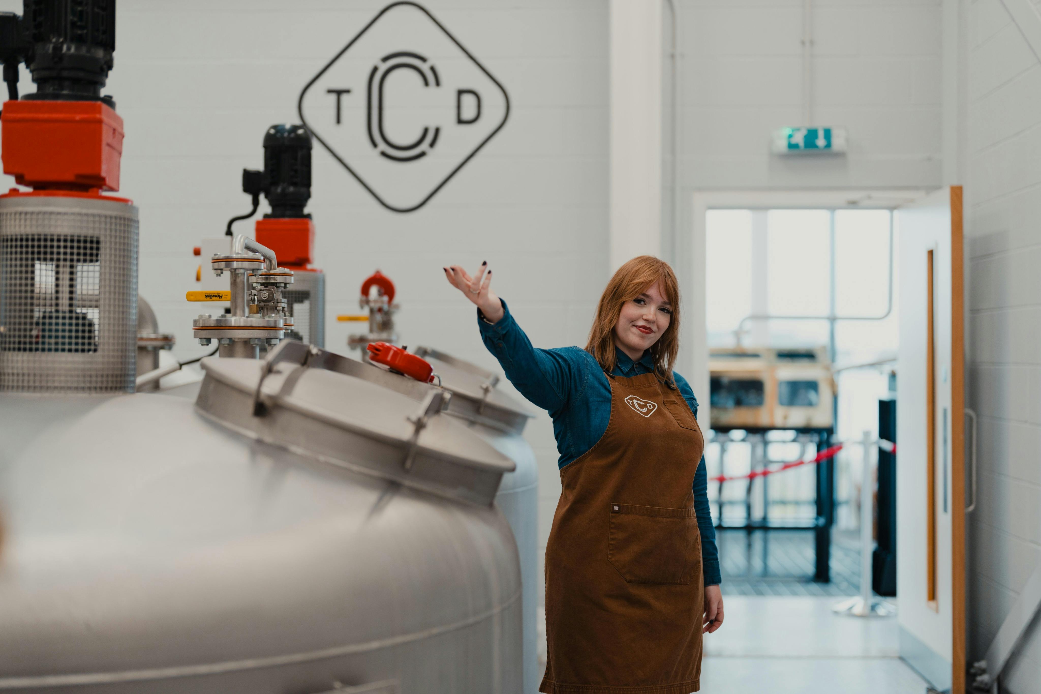 En kvinde i et brunt forklæde står i et destilleri, smilende og gestikulerende, med udstyr og et logo synligt i baggrunden.