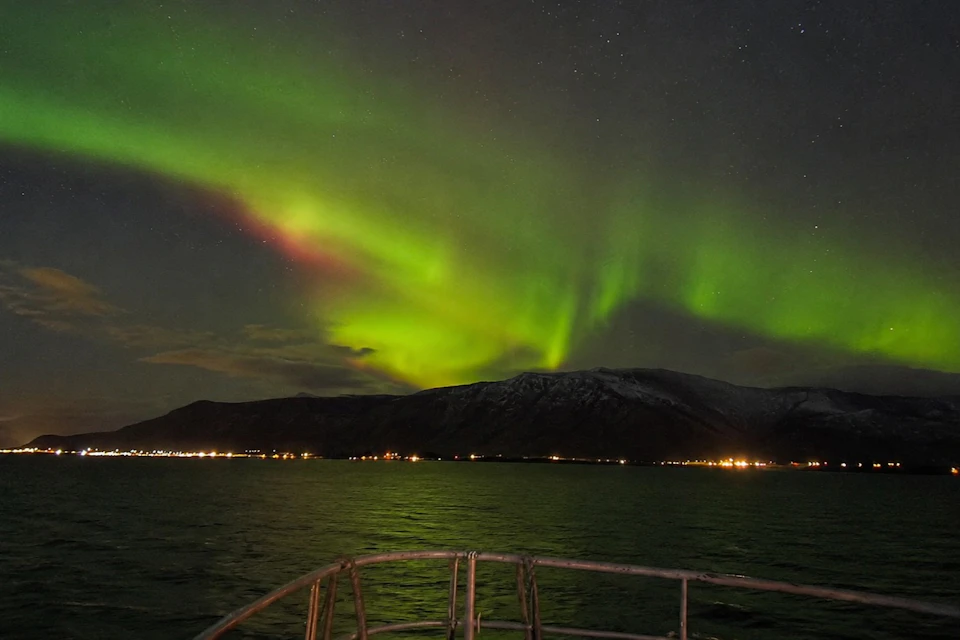 Aurora borealis with vibrant green and red hues over a mountain range, seen from a boat on calm water at night.
