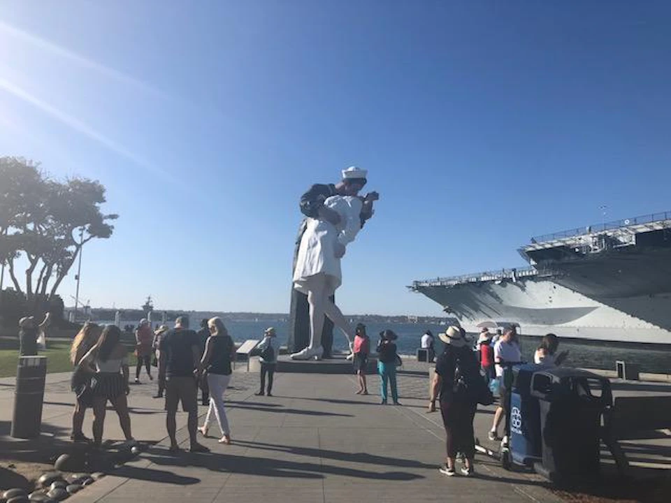 A large sculpture of a sailor kissing a nurse stands near a harbor, with people gathered around and a ship in the background.