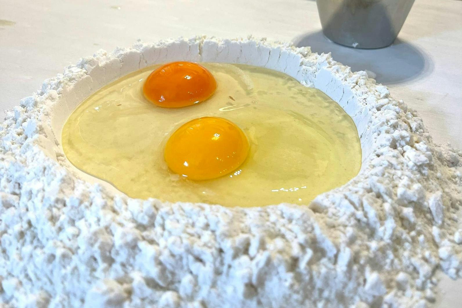 Two raw eggs cracked into a well of flour on a countertop, with a metal cup partially visible in the background.